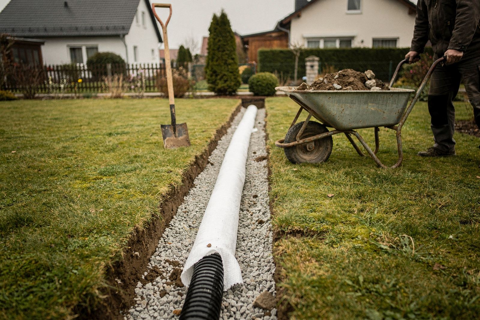 Offener Graben im Garten mit Drainagerohr auf Kies, seitlich eingeschlagenes Filtervlies